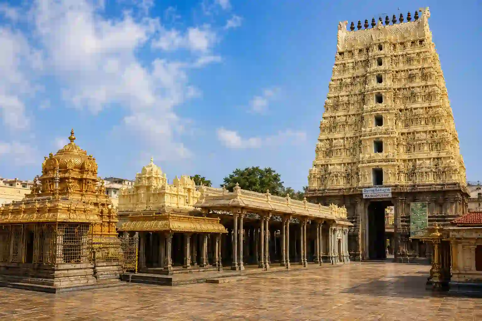 Ancient intricately carved Hindu temple complex with tall gopuram tower, golden shrines, and stone pillars under a partly cloudy sky.