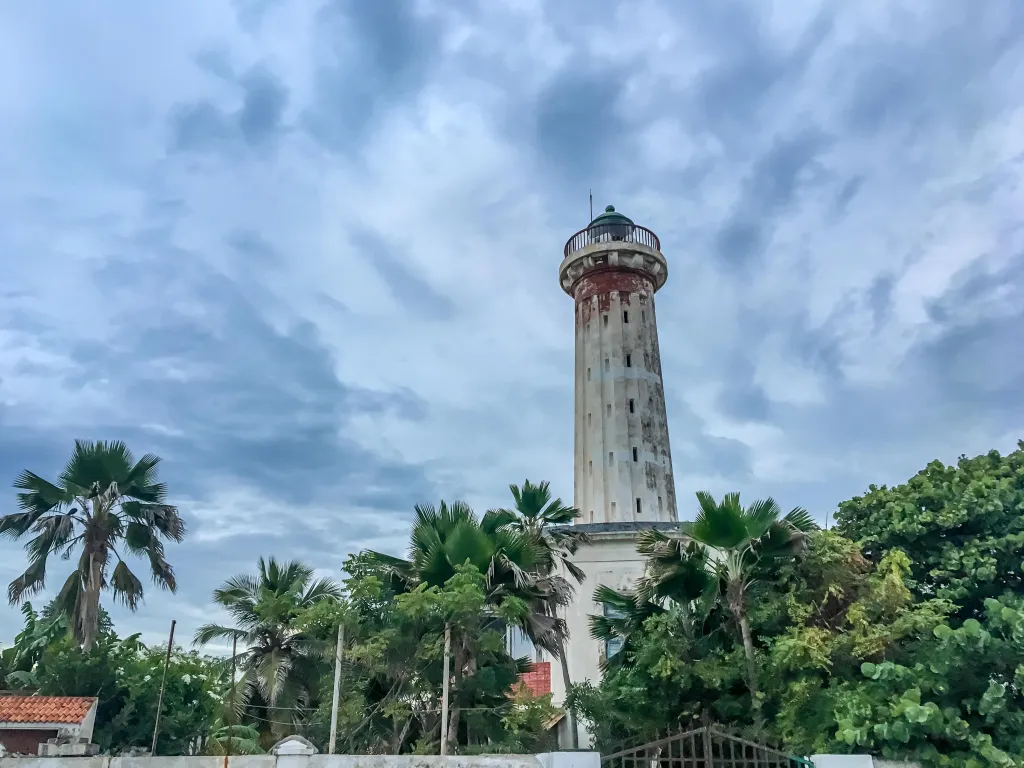 Tall weathered lighthouse surrounded by palm trees and greenery under a cloudy blue sky.