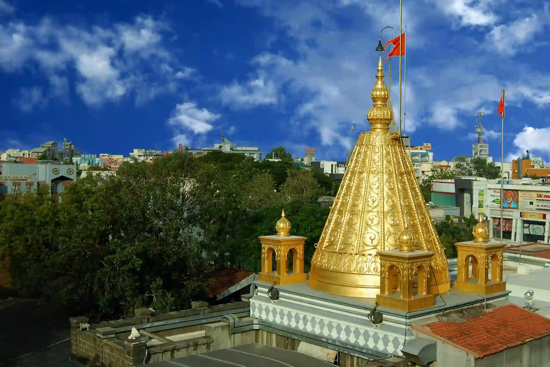Golden temple dome with intricately carved patterns, orange flags, surrounded by trees and city buildings under a blue sky with scattered clouds.