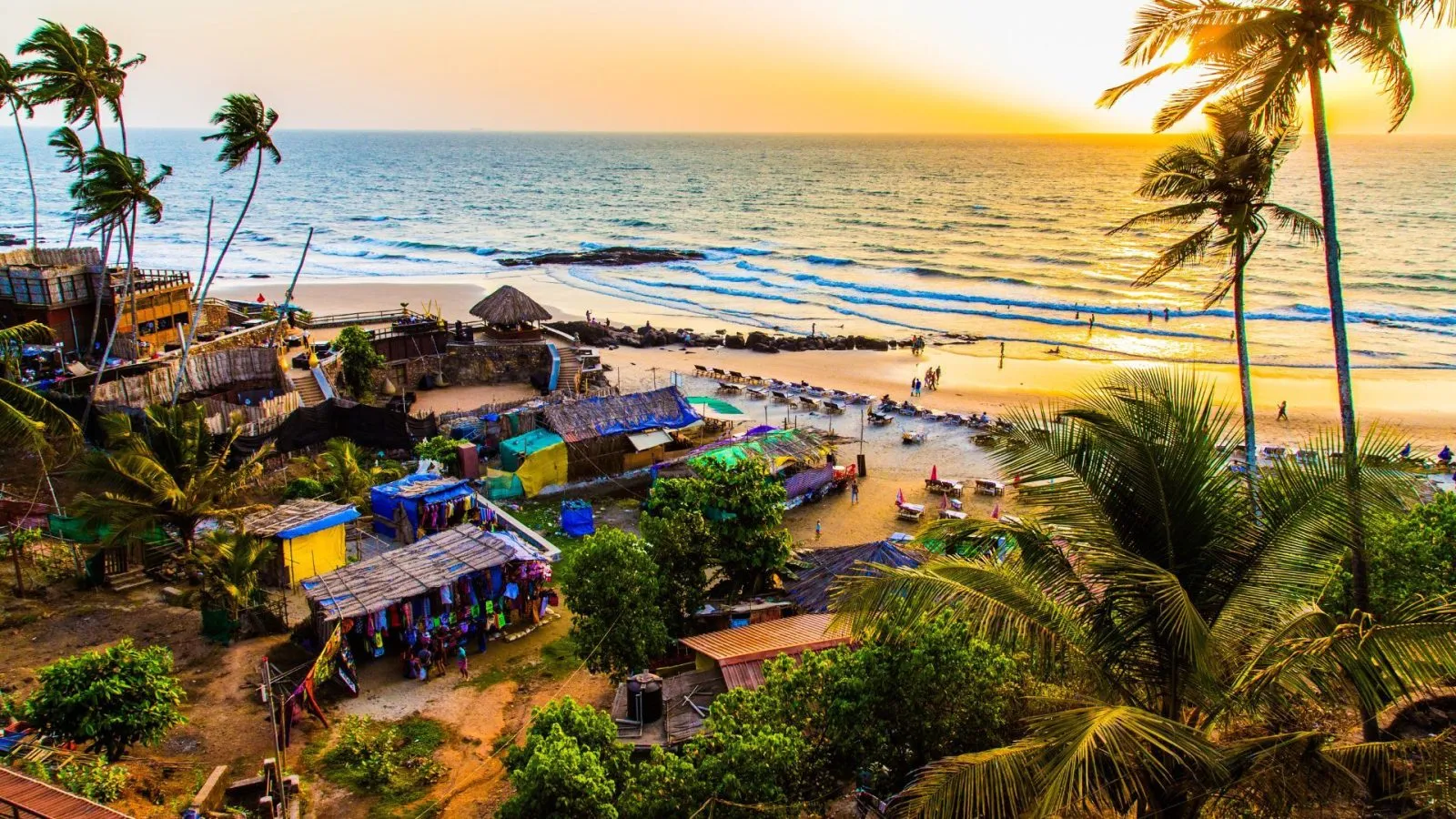 Sunset view of a tropical beach with palm trees, colorful huts, and people enjoying the shoreline and sea waves.
