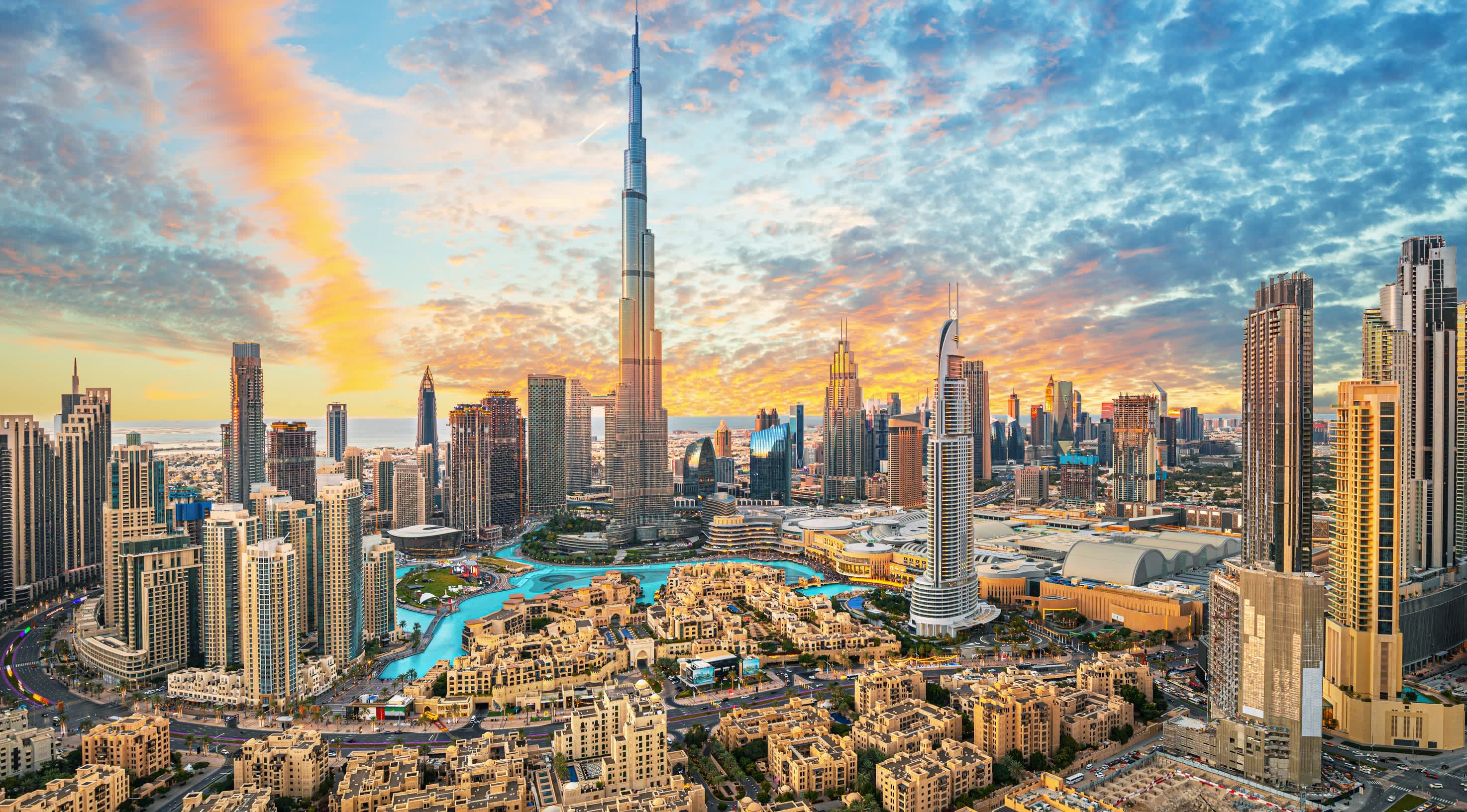 Panoramic view of Dubai skyline at sunset featuring Burj Khalifa, modern skyscrapers, blue waterways, and vibrant skies with scattered clouds.
