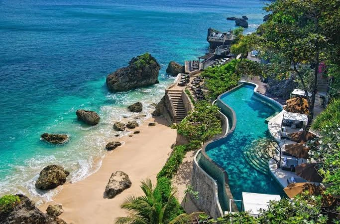 Oceanfront resort pool overlooking a sandy beach with rocks and turquoise water surrounded by tropical greenery.