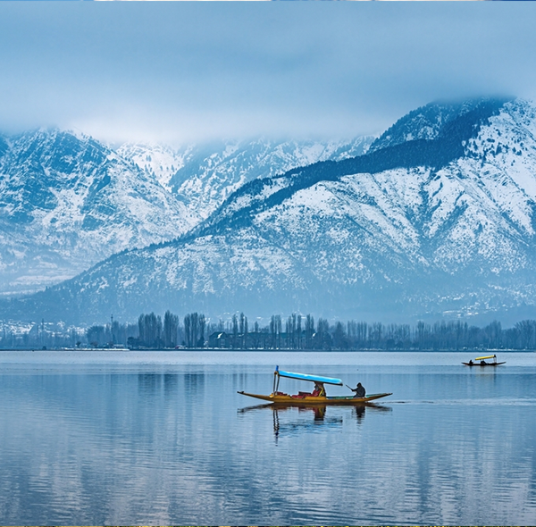 Calm lake with two boats and snow-covered mountains partly shrouded in clouds in the background.